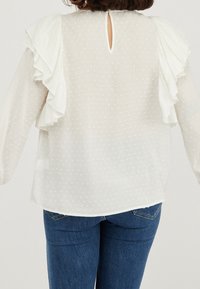 Valkoinen paita, jossa on tekstuurisia pilkkuja, röyhelöhihoja ja takaosan avainaukko. Yhdistetty sinisiin farkkuihin kontrastiksi.