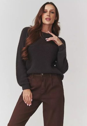 Kobieta z długimi brązowymi włosami, ubrana w czarny sweter i brązowe spodnie, pozuje z jedną ręką blisko twarzy, a drugą na udzie, na jednolitym tle.