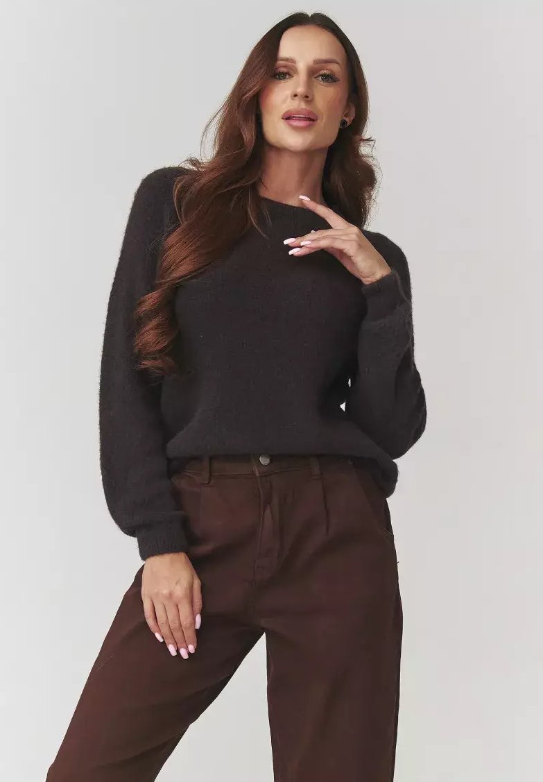 Kobieta z długimi brązowymi włosami, ubrana w czarny sweter i brązowe spodnie, pozuje z jedną ręką blisko twarzy, a drugą na udzie, na jednolitym tle.