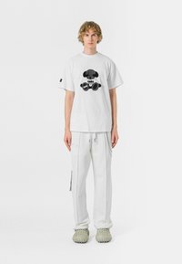 Camiseta blanca de algodón con un diseño gr�áfico negro, combinada con pantalones deportivos blancos con un detalle de cremallera y zapatos gris claro.