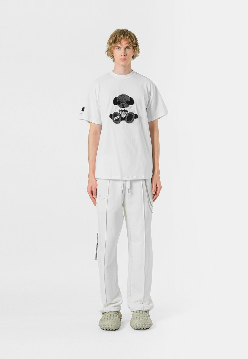 Camiseta blanca de algodón con un diseño gráfico negro, combinada con pantalones deportivos blancos con un detalle de cremallera y zapatos gris claro.