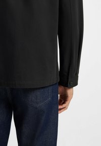Sort jakke med en glat tekstur, knappet manchet og lige kant, parret med mørkeblå denim jeans med sømme på baglommerne.