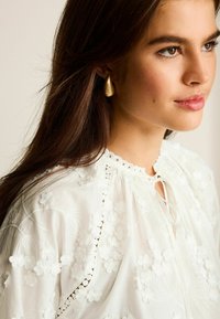 Biela vyšívaná blúzka s kvetinovými vzormi, krajkovým lemovaním a uzatváraním na kľúčovú dierku. Modelka má na sebe zlaté slzičkové náušnice.