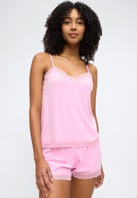 Γυναίκα με σγουρά μαλλιά που φοράει ροζ καλοκαιρινό τοπ με δαντέλα και αντίστοιχο σορτς, στέκεται μπροστά σε φόντο μονόχρωμο.