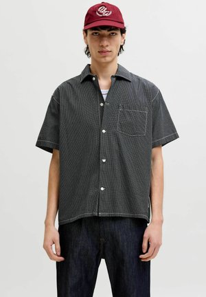 Jeune homme portant une chemise à manches courtes à carreaux noir et blanc, un jean foncé et une casquette de baseball rouge, debout devant un fond uni.