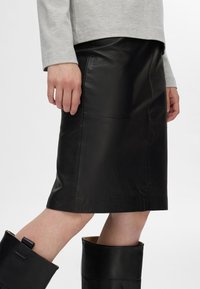 Zwarte leren kokerrok met een gladde textuur, op knielengte, met zijnaad en een getailleerde silhouette. Draagt met hoge zwarte laarzen.