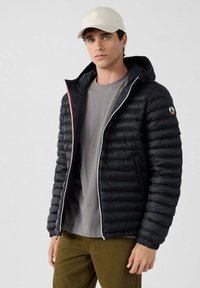 Piumino nero con cappuccio, caratterizzato da cuciture orizzontali, una cerniera argentata e dettagli rossi. Indossato sopra una maglietta grigia e pantaloni oliva.