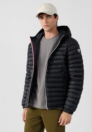 Zwarte pufferjack met een capuchon, voorzien van horizontale stiknaden, een zilveren rits en rode accenten. Draag dit over een grijze shirt en olijfgroene broek.