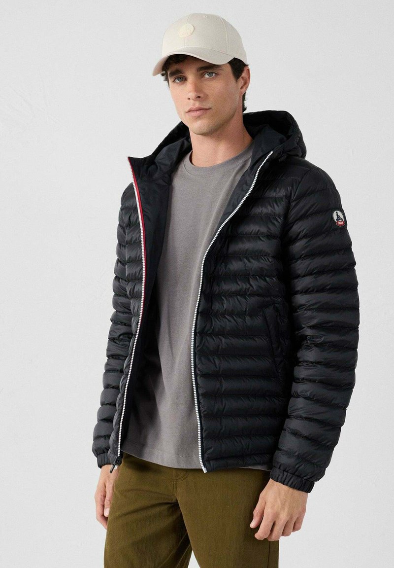 Piumino nero con cappuccio, caratterizzato da cuciture orizzontali, una cerniera argentata e dettagli rossi. Indossato sopra una maglietta grigia e pantaloni oliva.