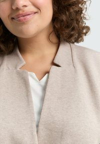 Chaqueta de punto beige con un acabado texturizado, que presenta un sutil patrón de rayas y un escote en forma de V sobre una blusa blanca.