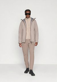 Bėgių atspari smėlio spalvos striukė su pilkais akcentais, derinama su smėlio spalvos vidiniu sluoksniu ir derančiomis kelnėmis. Juodi batai ir akiniai nuo saulės papildo išvaizdą.