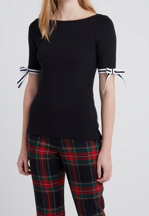 Vrouw draagt een zwarte top met korte mouwen met witte gestreepte strikjes op de mouwen en roodgroene geruite broek, staand tegen een effen achtergrond.