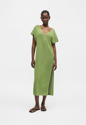 Woman wearing a green knit midi dress with V-neck, short sleeves, side slit, necklace, earrings, and brown flat sandals, standing on white background.