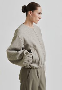 Grå, teksturert hettegenser med rund hals, glidelåslukking og voluminøse ermer. Parret med løse, olivengrønne bukser.