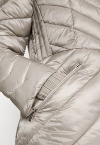 Jasnobeżowa kurtka puchowa z pikowanym wzorem, z zamkniętą kieszenią i ściągaczem z dzianiny widocznym na rękawie. Miękka, błyszcząca tekstura.