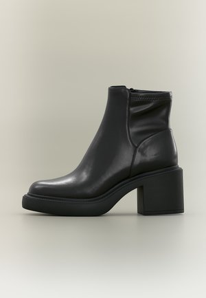 Bottine en cuir noir avec un talon bloc épais et une fermeture éclair latérale sur un fond neutre clair.