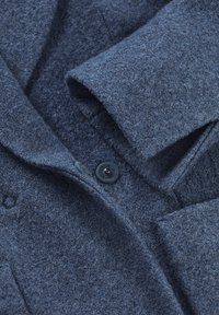 Mörkblå ullrock med en mjuk textur, försedd med en synlig svart knapp och vikt krage. Detaljer inkluderar en slät innerfoder.