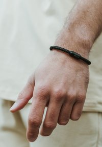 Pulsera trenzada negra con un broche liso, llevada en una mano que muestra una piel texturada; el fondo presenta una tela neutra.