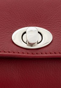 Rdeča usnjena torbica v zoomu z ovalnim srebrnim zapiranjem, zapletenim šivanjem in gladko teksturo na površini.