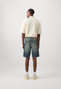 Polo bej cu mânecă scurtă, cu un model texturat, asociat cu pantaloni scurți din denim albastru estompat. Ciorapi albi și adidași de culoare deschisă completează ținuta.