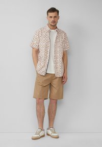 Kurzärmliges, knöpfbares Hemd in Hellpink mit braunen Blättermustern, kombiniert mit beigen Shorts und neutralen Sneakers. Glatte Stoffstruktur.