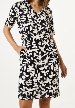 Femme portant une robe noire mi-longue à manches courtes avec un motif abstrait de feuilles blanches, les mains dans les poches latérales, un bracelet doré au poignet droit.