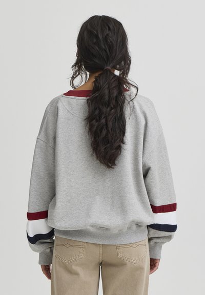 Sweat-shirt gris avec une coupe décontractée, présentant des accents à rayures bordeaux et marine sur les manches et un col côtelé. Associé à un pantalon beige.