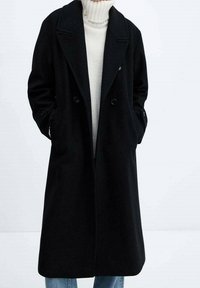 Cappotto lungo nero con design doppiopetto, ampi rever, tasche laterali e una texture liscia, indossato sopra un maglione chiaro.