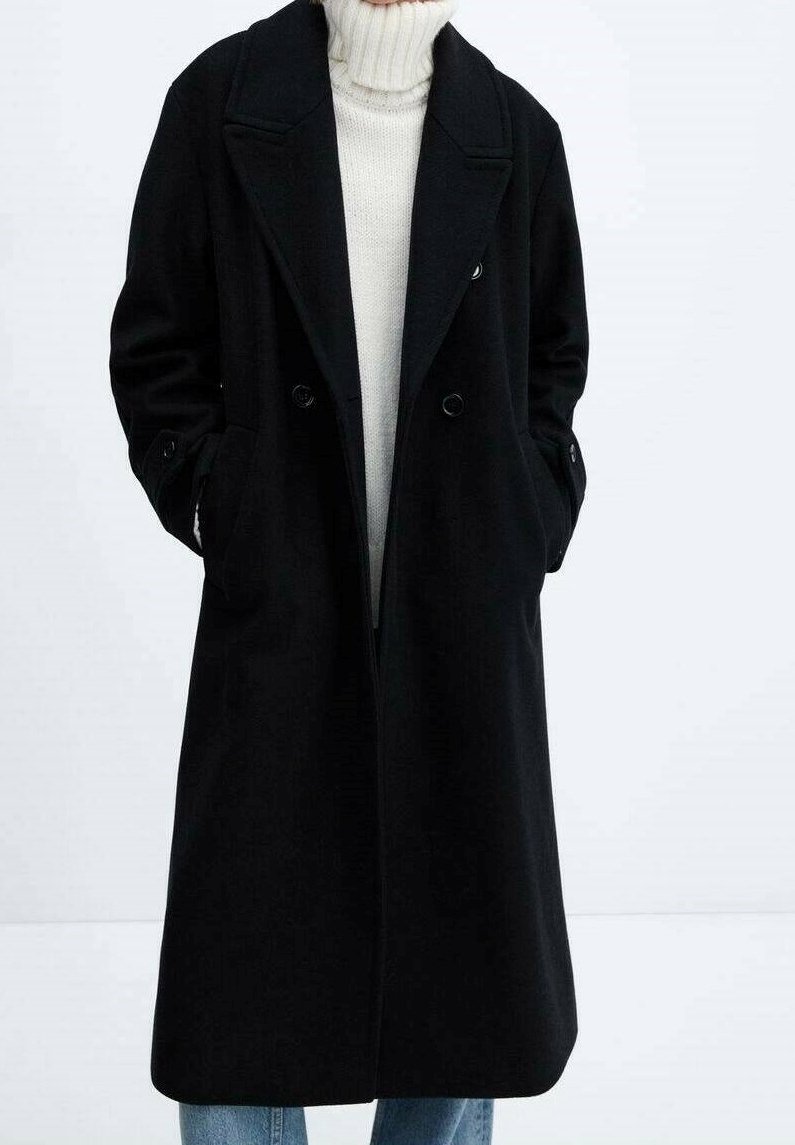 Cappotto lungo nero con design doppiopetto, ampi rever, tasche laterali e una texture liscia, indossato sopra un maglione chiaro.