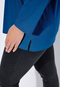 Niebieska bluzka z długim rękawem, okrągłym dekoltem i bocznymi rozcięciami, zestawiona z ciemnymi obcisłymi dżinsami. Tkanina wydaje się gładka i lekko teksturowana.