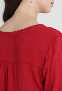 Blusa rossa con texture a costine, caratterizzata da un ampio scollo e una vestibilità morbida, progettata con una cucitura centrale e dettaglio arricciato sul retro.