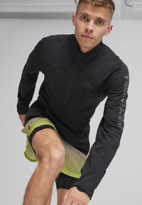 Musta urheilullinen puolihihainen paita, jossa on tekstuuroitu kangas ja pitkät hihansuut. Khakiväriset shortsit, joissa on neonvihreät yksityiskohdat ja verkkokangas. Säädettävä kaulus.