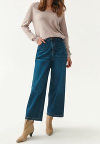 Jeansy z wysokim stanem i szerokimi nogawkami w ciemnoniebieskim denimu, zestawione z jasnobeżową bluzką z długim rękawem oraz beżowymi botkami na kostkę z ostrym noskiem.