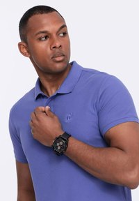Camisa polo morada con un logotipo de marca en el pecho, combinada con un reloj de pulsera negro con esfera redonda y múltiples diales.