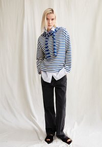 Blau-weiß gestreifter, übergroßer Pullover mit einer Bindung, über einem weißen Hemd getragen, kombiniert mit schwarzen, weit geschnittenen Hosen und schwarzen Sandalen.