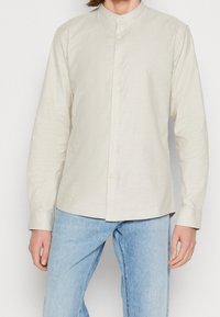 Helle beige Langarmbluse mit Stehkragen, Knopfverschluss vorne und einer glatten Textur, kombiniert mit hellblauen Jeans.