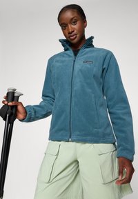 Veste en polaire teal, avec fermeture éclair et col montant, ornée d'un patch logo. Associée à un short cargo vert clair et des bâtons de randonnée.