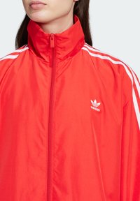 Punainen vetoketjullinen takki, joka on valmistettu kevyestä materiaalista. Takissa on valkoiset raidat olkapäiden kohdalla ja vasemmassa rinnassa kirjailtu logo.