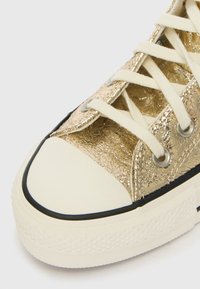 Sapatilha metálica dourada com superfície texturizada, atacadores em tom creme e biqueira em borracha branca. Apresenta um acabamento preto ao longo da sola e dos ilhós.