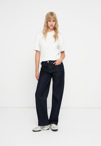 Lengvas baltas marškinėliai, tamsiai mėlynos plačios kelnės ir pilki batai. Kelnės turi aukštą liemeni ir klasikines kišenes su metaliniais akcentais.