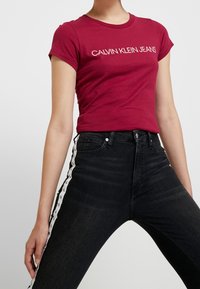 Camiseta de algodón roja con "CALVIN KLEIN JEANS" impreso en blanco. Llevada con jeans negros de cintura alta con rayas de logo blancas en los laterales.