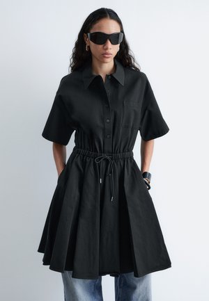 Donna con vestito nero a maniche corte con bottoni e vita stretta, grandi occhiali da sole neri, orecchini a cerchio, braccialetti e jeans blu.