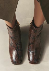 Pruunid ja mustad kääriku prindiga pahkluu saapad, millel on läikiv viimistlus, ruudukujuline nina, külgmised tõmblukud ja peened tekstuurilised detailid.