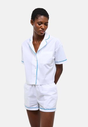 Femme portant une chemise blanche à manches courtes boutonnée et un short assorti avec un bord festonné bleu, debout devant un fond clair uni.