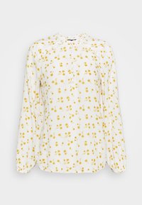 Blouse blanche en tissu léger, avec un motif floral jaune, devant boutonné et manches longues élastiquées. Détail de col volanté.