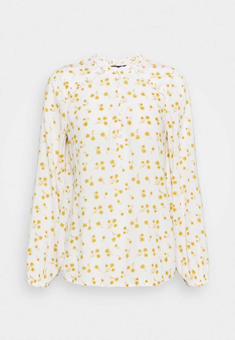 Blouse blanche en tissu léger, avec un motif floral jaune, devant boutonné et manches longues élastiquées. Détail de col volanté.