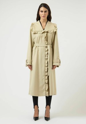 Kvinde iført beige trenchcoat med bælte, flæsekrave og front, sorte bukser og sorte spidse hæle, stående foran en hvid baggrund.