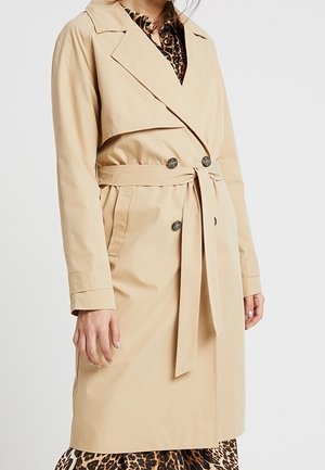 Beige dobbeltradet trenchcoat med bælte bundet i taljen, båret over en leopardprint-kjole, vist fra skuldre til knæ.