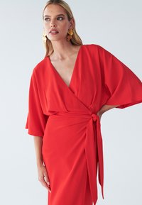 Rotes Wickelkleid mit kurzen, schwingenden Ärmeln, tiefem V-Ausschnitt und seitlichem Knoten-Detail. Glattes, leichtes Material mit fließender Silhouette.