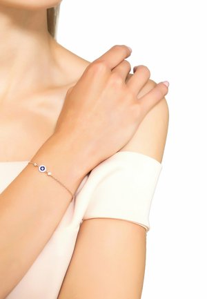 Guldfarvet armbånd med et blåt øje-motiv, små klare ædelstene og en delikat kæde, båret på en lys hudarm. Glat hudstruktur.